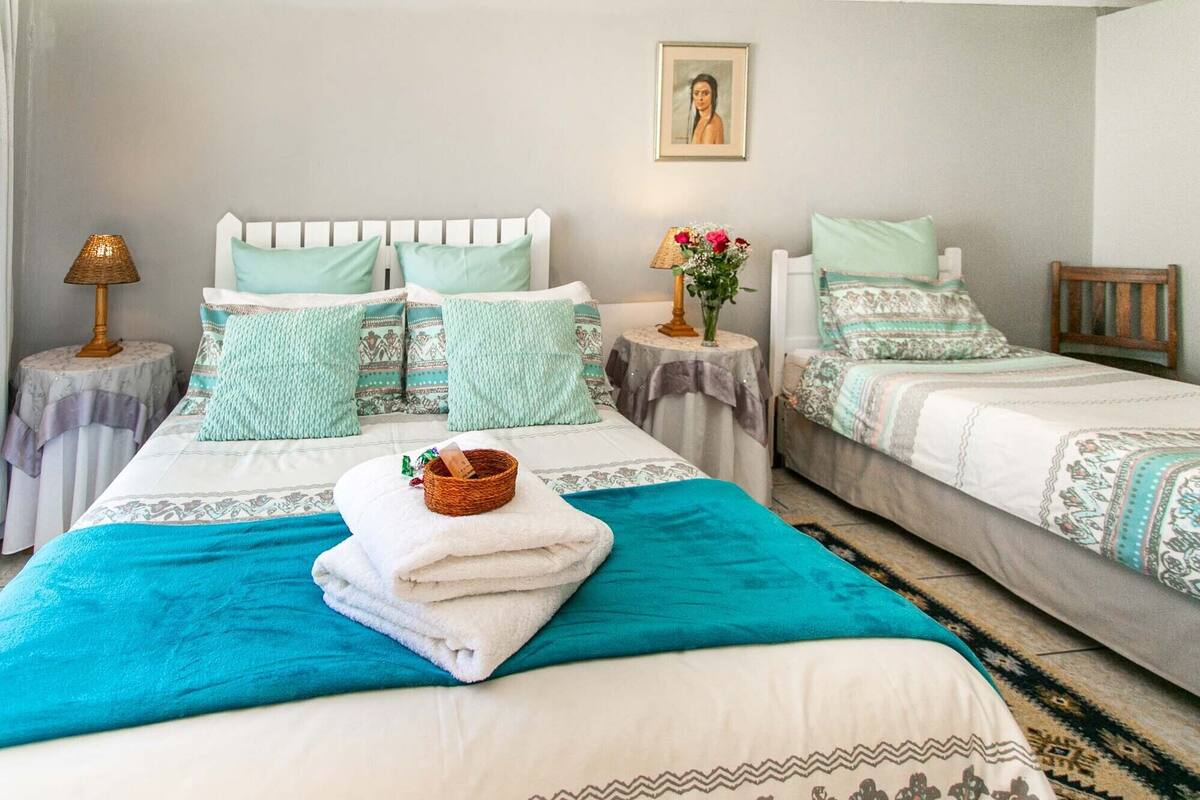 Une chambre d’hôtel avec deux lits simples, un grand lit recouvert d’une couverture turquoise, et une petite table avec une lampe et un vase de fleurs.