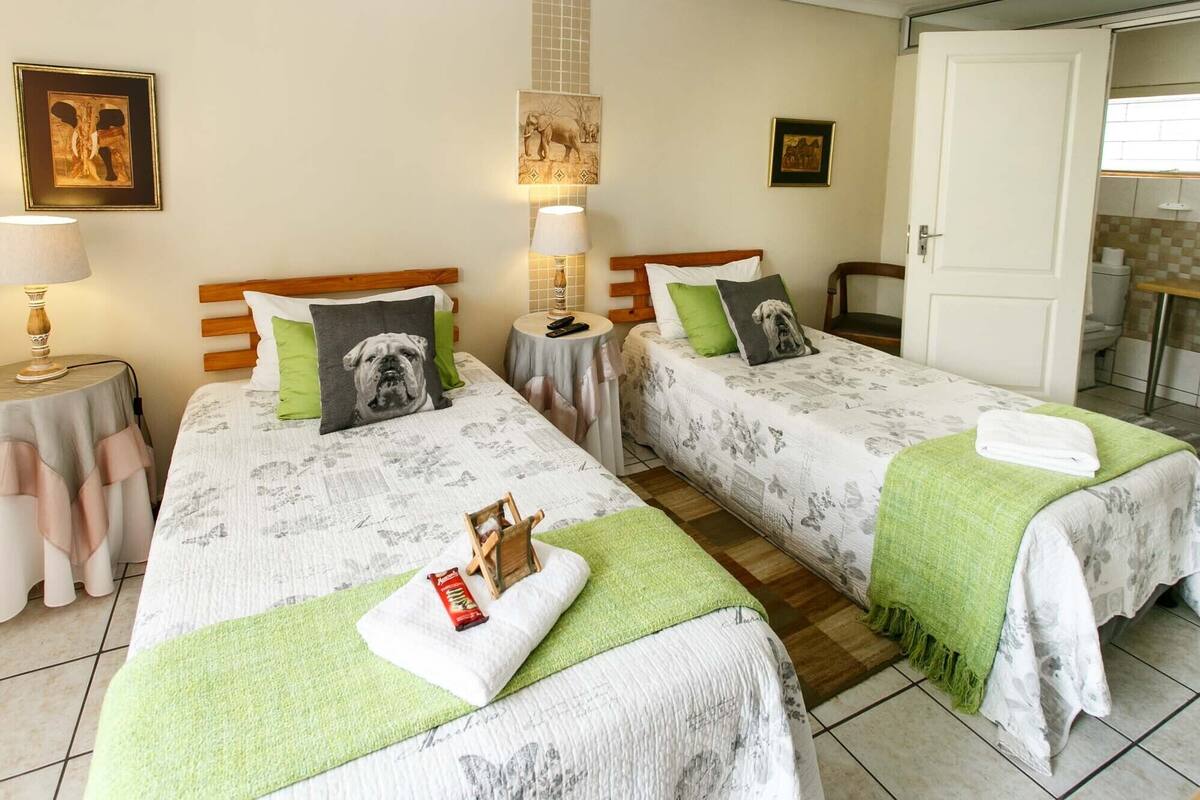 Une chambre d’hôtel avec deux lits, chacun recouvert d’une couvre-lit à motifs floraux et agrémenté d’un oreiller orné d’un motif de chien. On y trouve deux tables de chevet avec des lampes, une petite table ronde avec une lampe, et une porte donnant sur une salle de bain.
