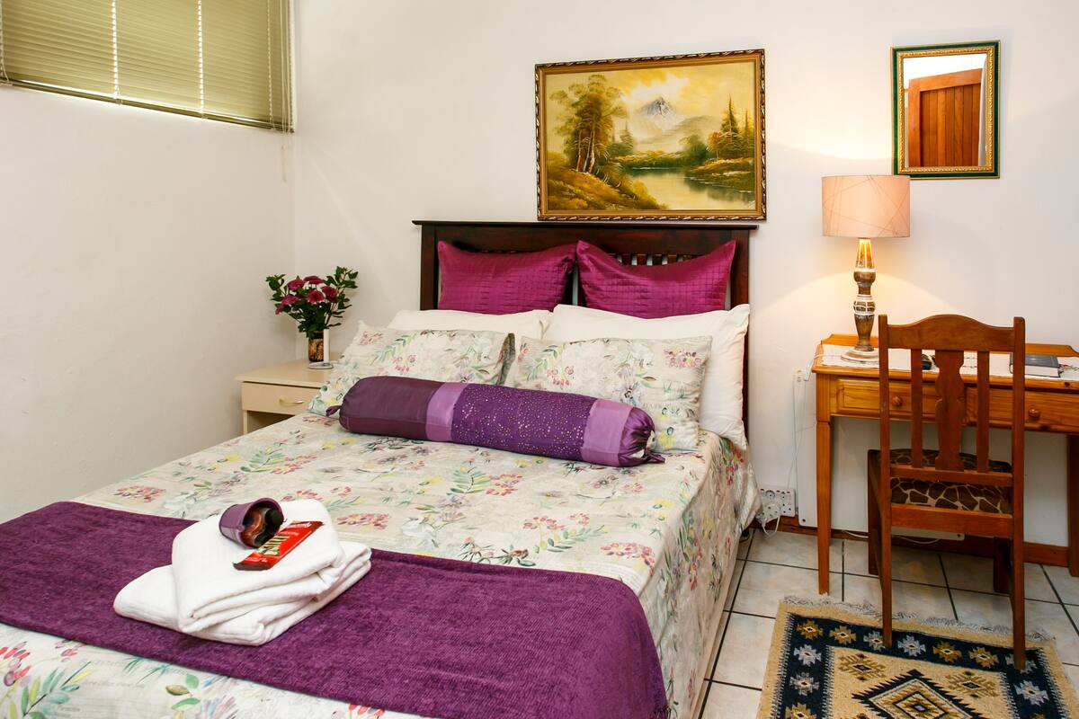 Un lit bien fait, recouvert d’une couvre-lit à motifs floraux, avec un oreiller violet et une serviette blanche sur laquelle repose une tablette de chocolat. Un bureau et une chaise en bois sont placés à côté du lit.