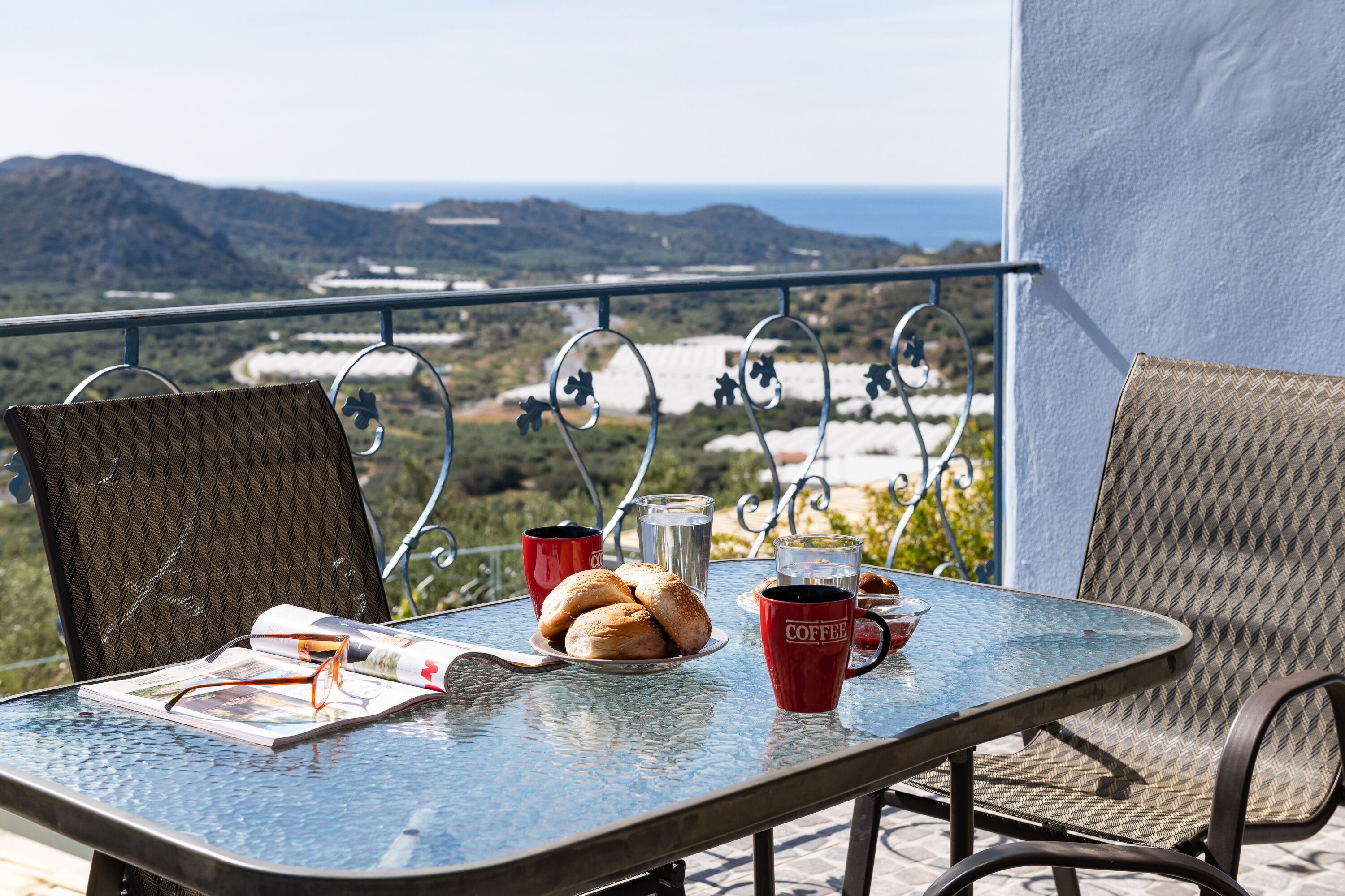Une table avec un magazine, des lunettes, une assiette de croissants et des tasses de café, le tout placé sur un balcon offrant une vue panoramique sur des collines et la mer.