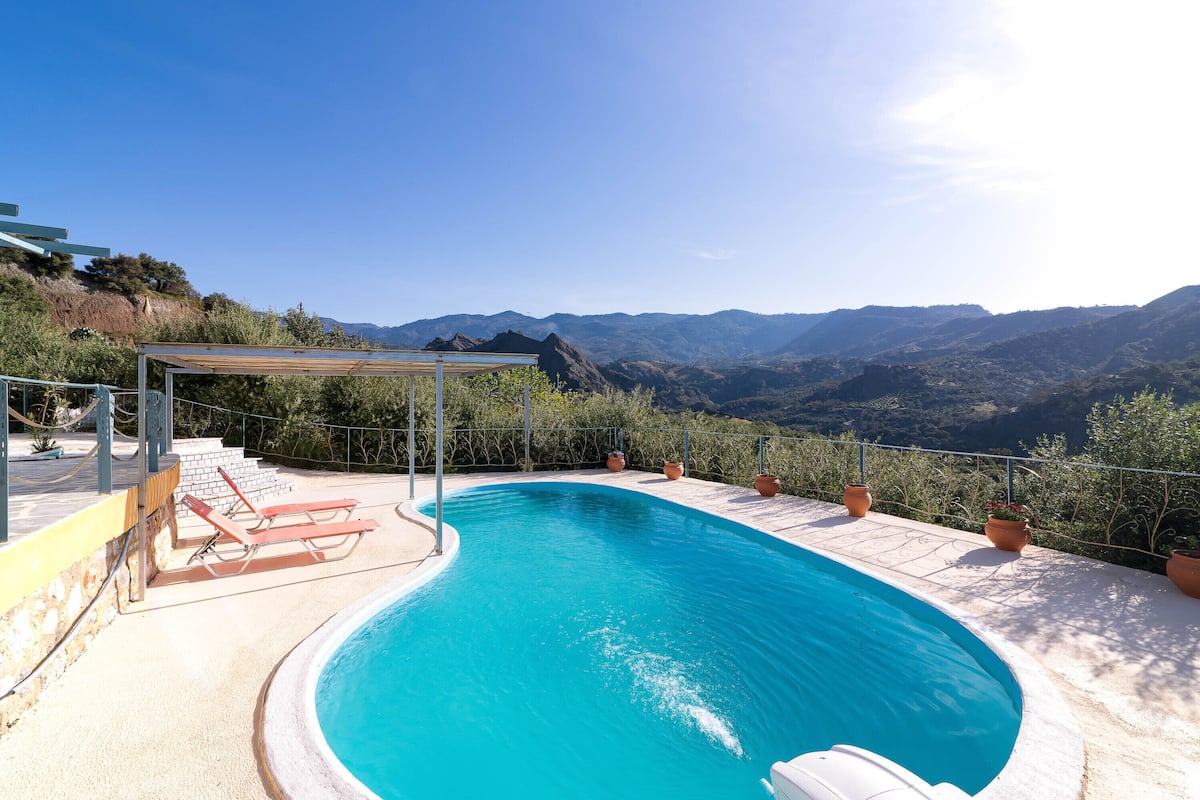 A swimming pool with a view of mountains, a covered seating area, and potted plants.