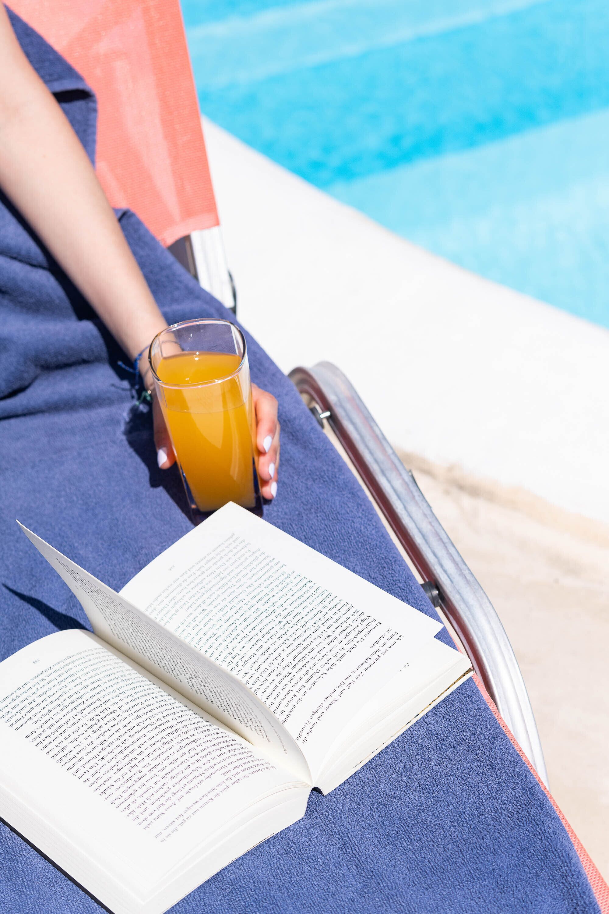 A person reclining on a blue lounge chair by a pool, holding a glass of orange juice, with an open book in front.