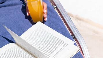 A person reclining on a blue lounge chair by a pool, holding a glass of orange juice, with an open book in front.