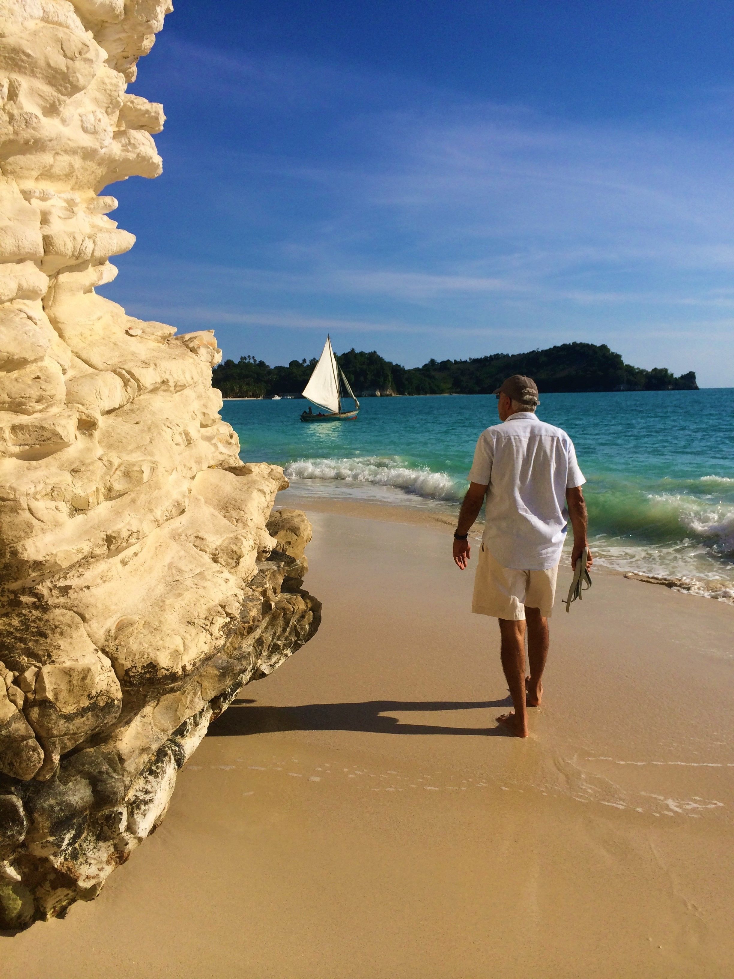A man walking on a sandy beach with a sailboat in the background.