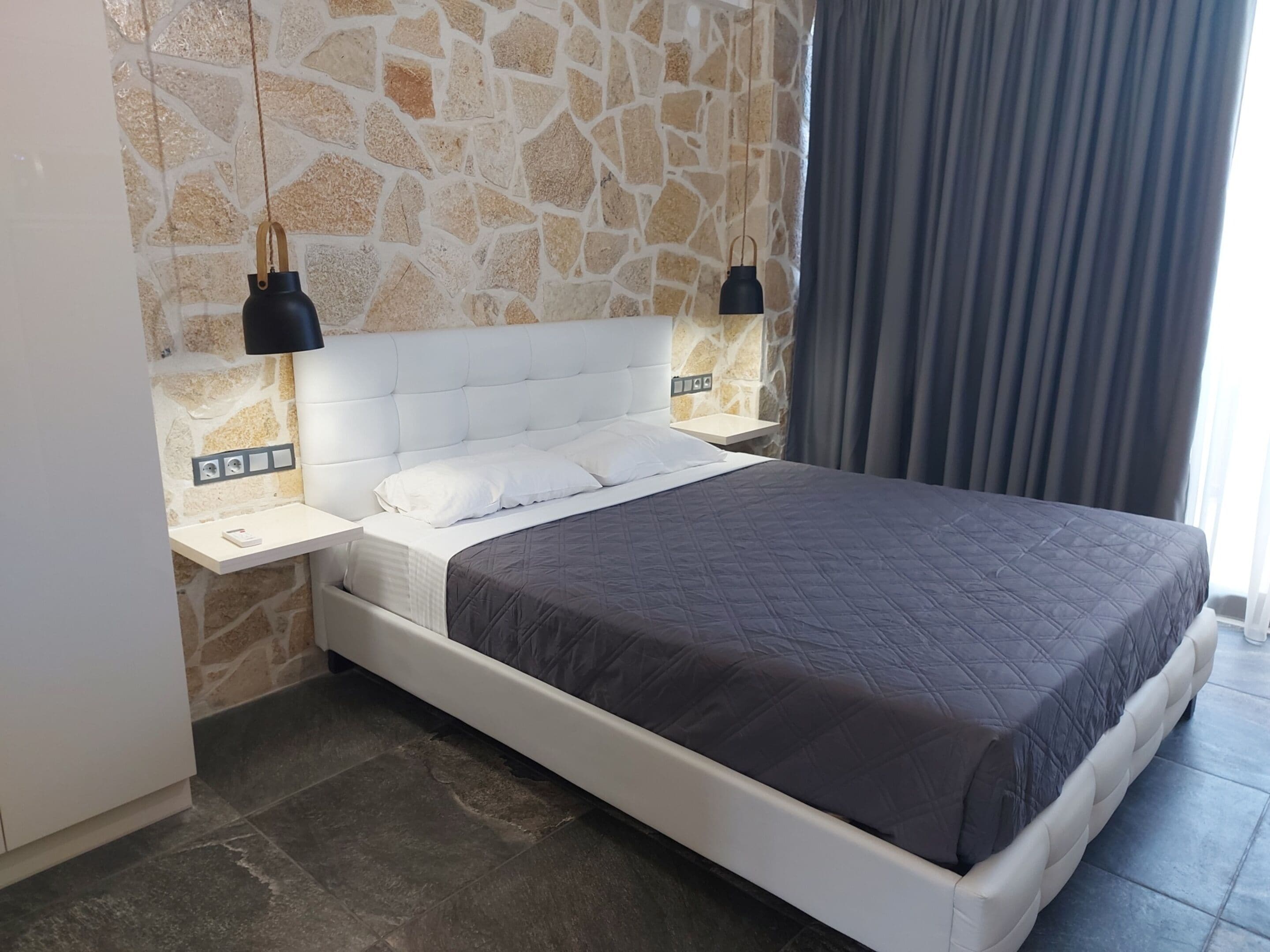 A modern bedroom with a stone wall, a white headboard, a dark quilted bedspread, and two black pendant lights.