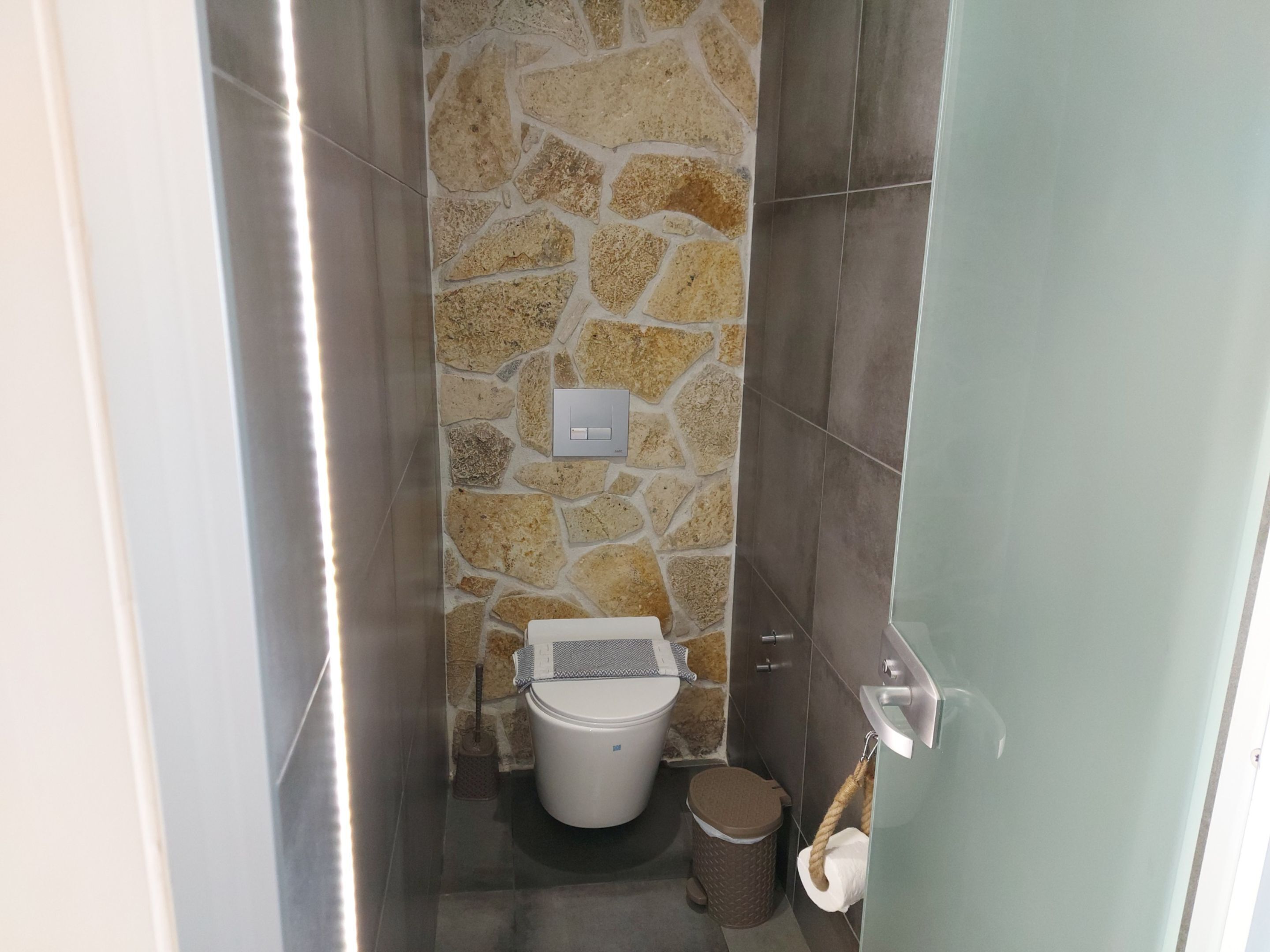 A small bathroom with a stone wall, a toilet, a trash bin, and a towel rack.