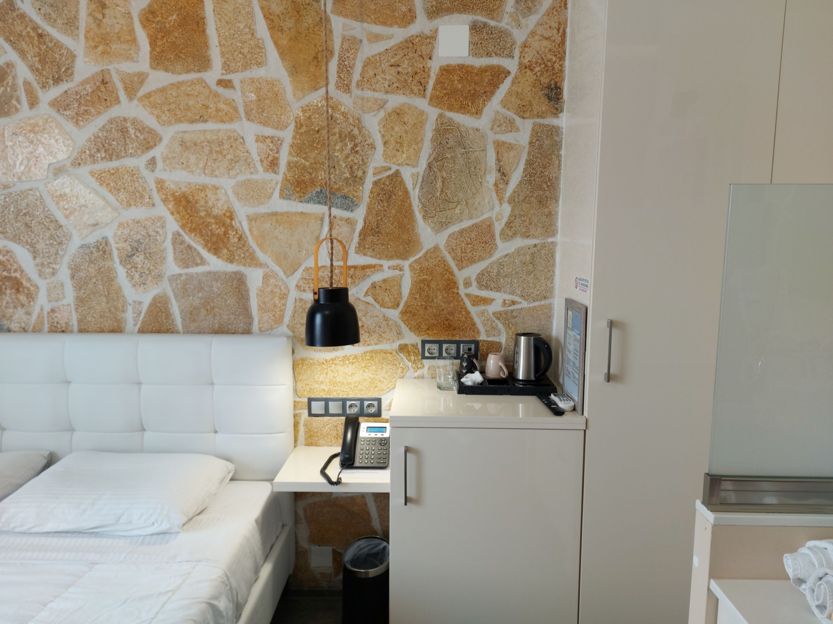 A hotel room with a stone wall, a bed, a bedside table with a phone, a kettle, and a small cabinet.