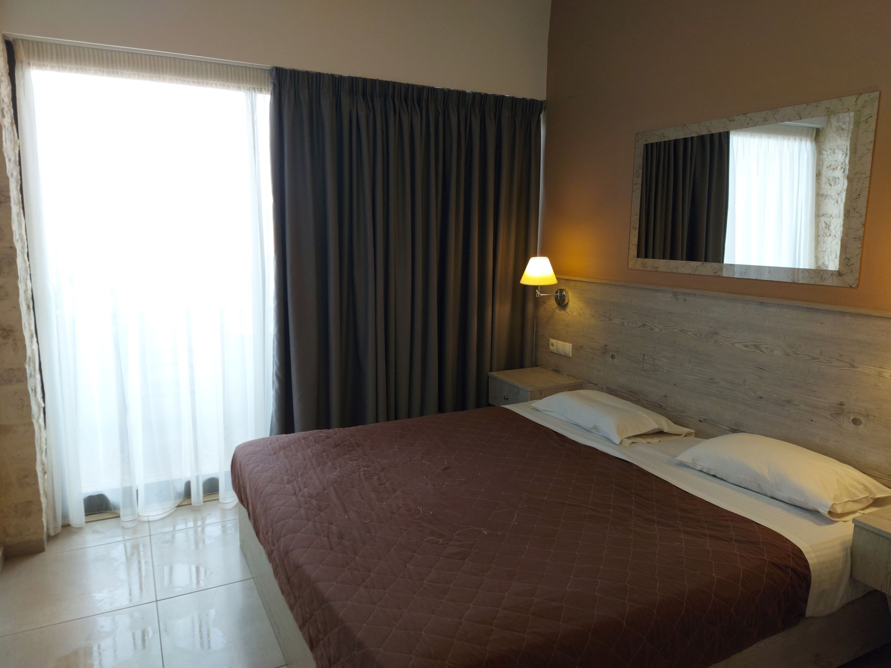 A hotel room with a large bed, a nightstand, a wall-mounted lamp, and a window with curtains.