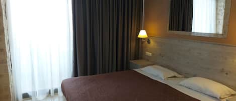 A hotel room with a large bed, a nightstand, a wall-mounted lamp, and a window with curtains.