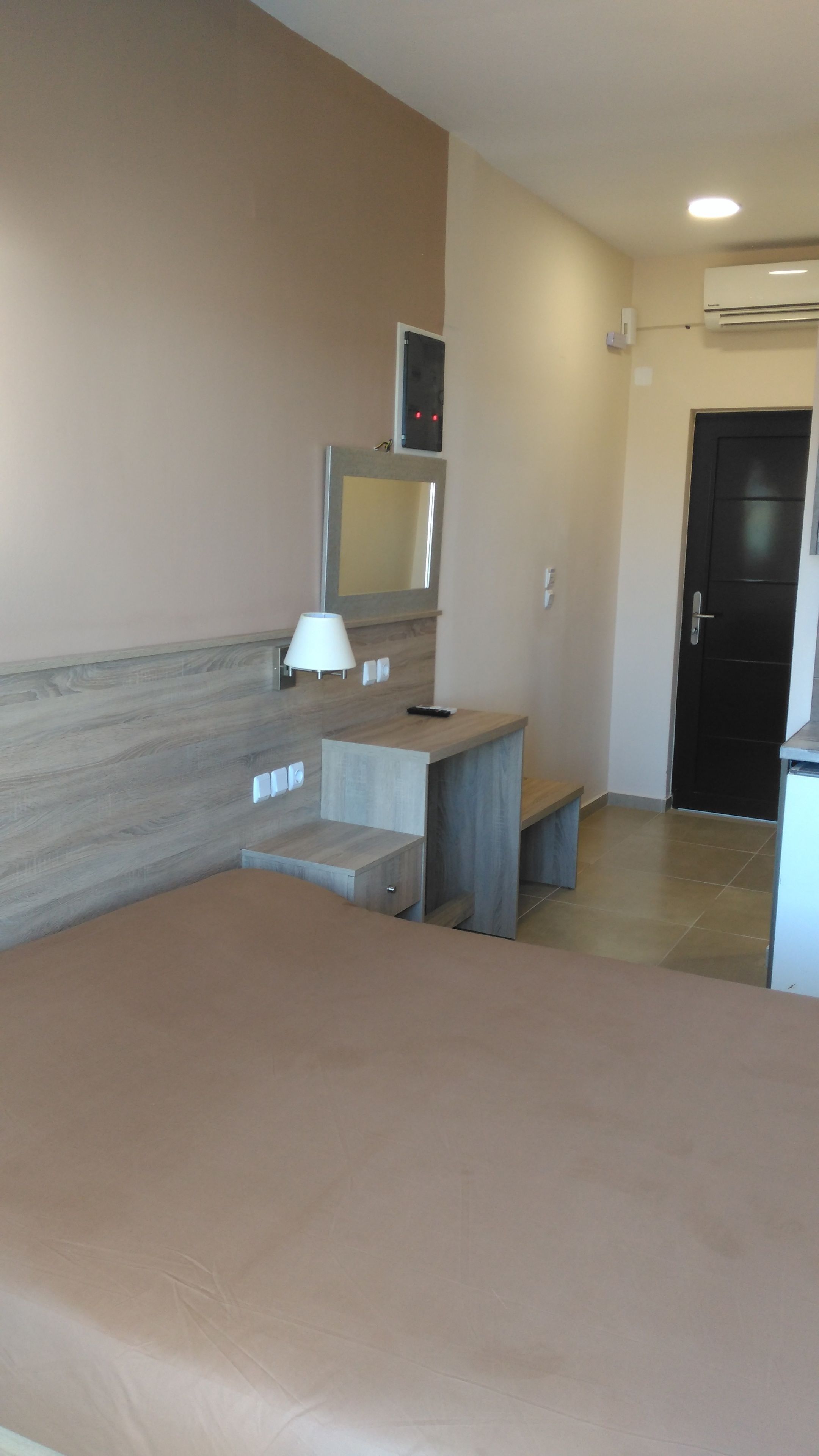 A hotel room with a bed, a desk with a lamp, a mirror, and a door.