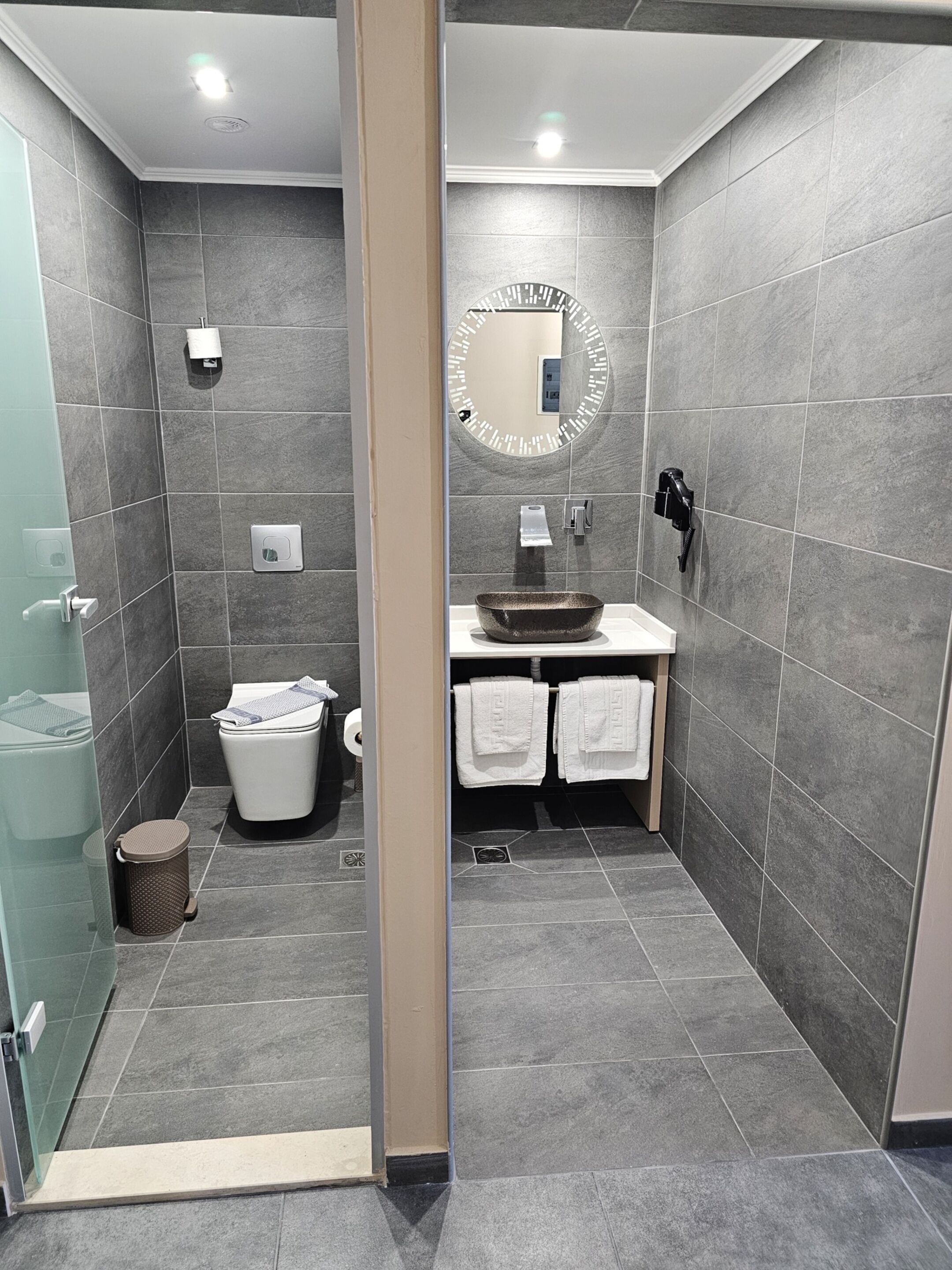 Une salle de bain moderne avec des carreaux gris, une douche en verre, un lavabo blanc et un miroir avec un cadre rond.