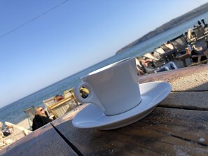 Eine weiße Tasse auf einem Holztisch mit Blick auf das Meer und Strandstühle im Hintergrund.