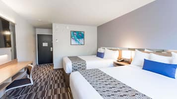 A hotel room with two beds, a desk, and a chair. There is a painting on the wall and a door in the background.