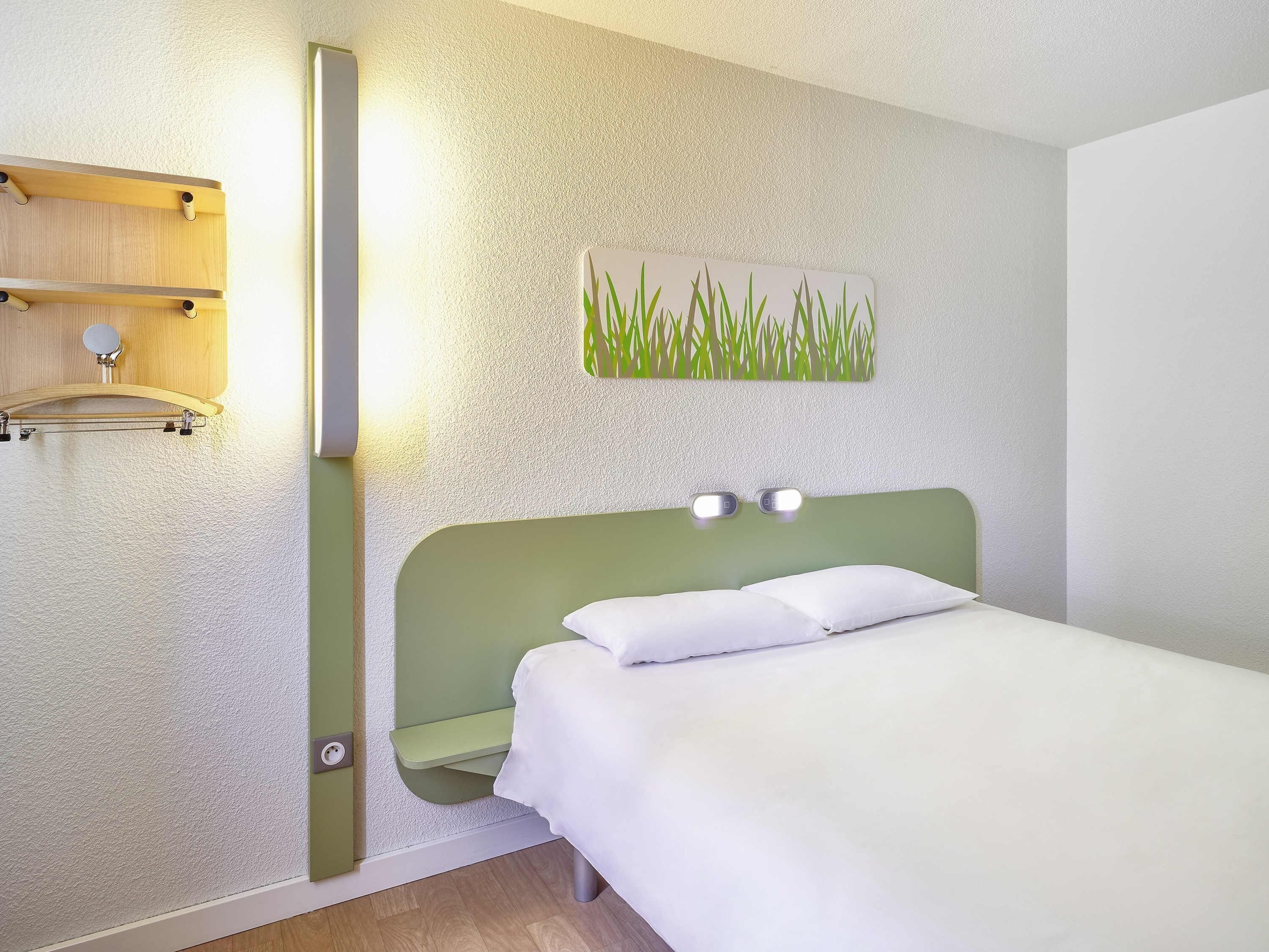 A hotel room with a bed, a wall-mounted shelf, a wall art piece, and a light fixture.