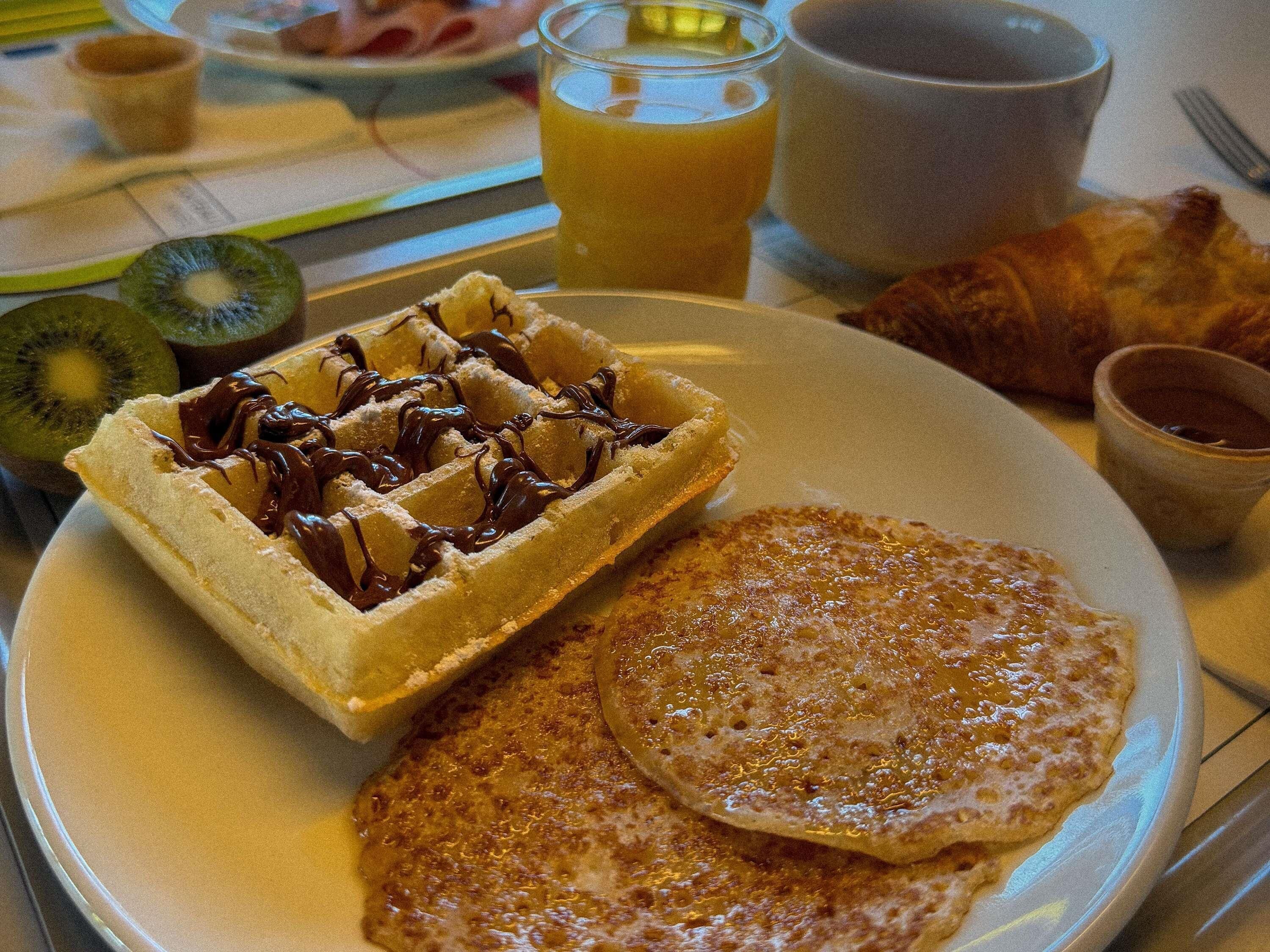 Une assiette avec des gaufres, des crêpes et un croissant, accompagnée d’un verre de jus d’orange et d’une tasse de café.