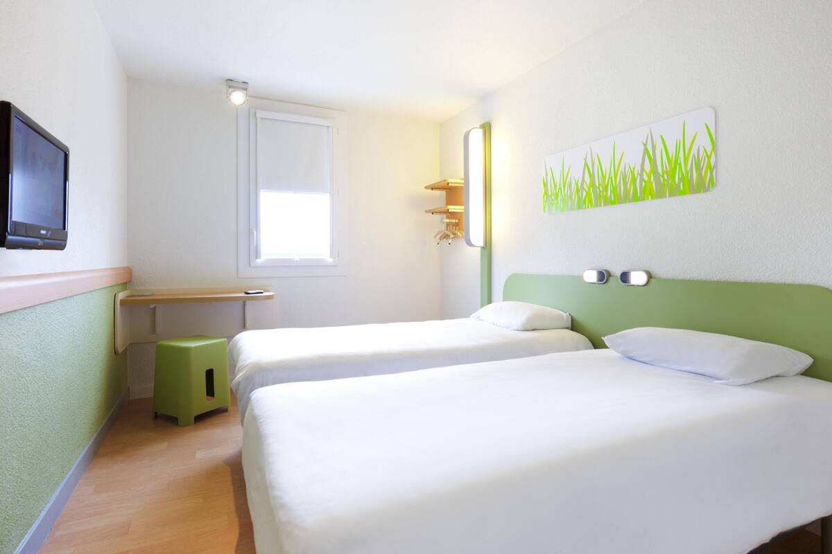 A hotel room with two beds, a desk, a small stool, a TV, a window, and a wall art piece.