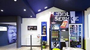 A modern interior with a reception desk, information kiosk, vending machines, and a seating area. - ibis Budget Dax Saint Paul les Dax (Saint-Paul-les-Dax)