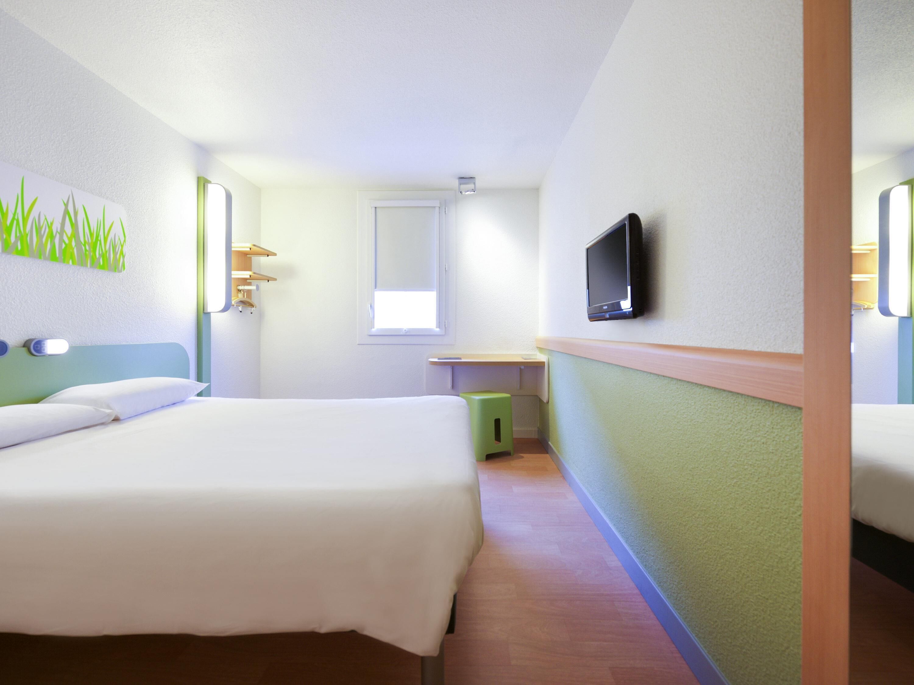 Habitación de hotel con dos camas, televisión, una ventana pequeña y un cesto de basura verde.