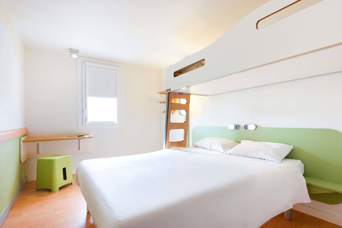 A hotel room with a bed, a small stool, a window, and a wall-mounted shelf.