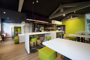 A modern bar area with a white counter, green stools, and a seating area with white tables and green ottomans. - ibis Budget Dax Saint Paul les Dax (Saint-Paul-les-Dax)