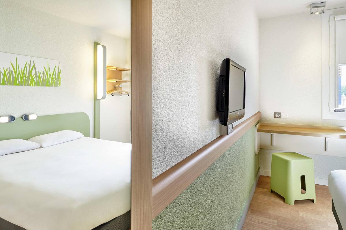 A hotel room with a bed, a television mounted on the wall, a small stool, and a window.