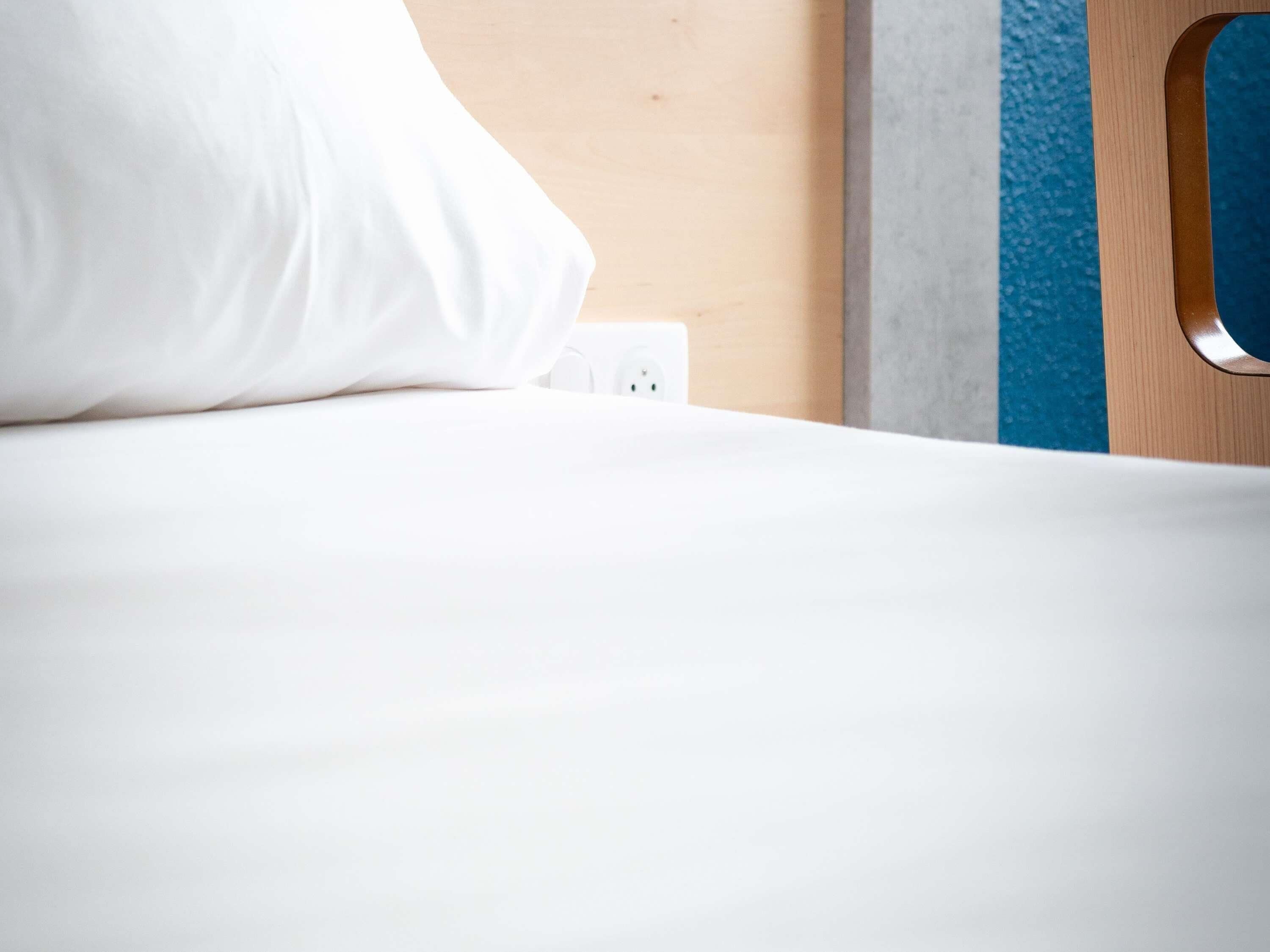 A neatly made bed with a white pillow and a power outlet on the wall.