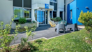Ein moderner Hotel Eingangsbereich mit blauer Beschilderung, Außenbestuhlung und viel Grün.