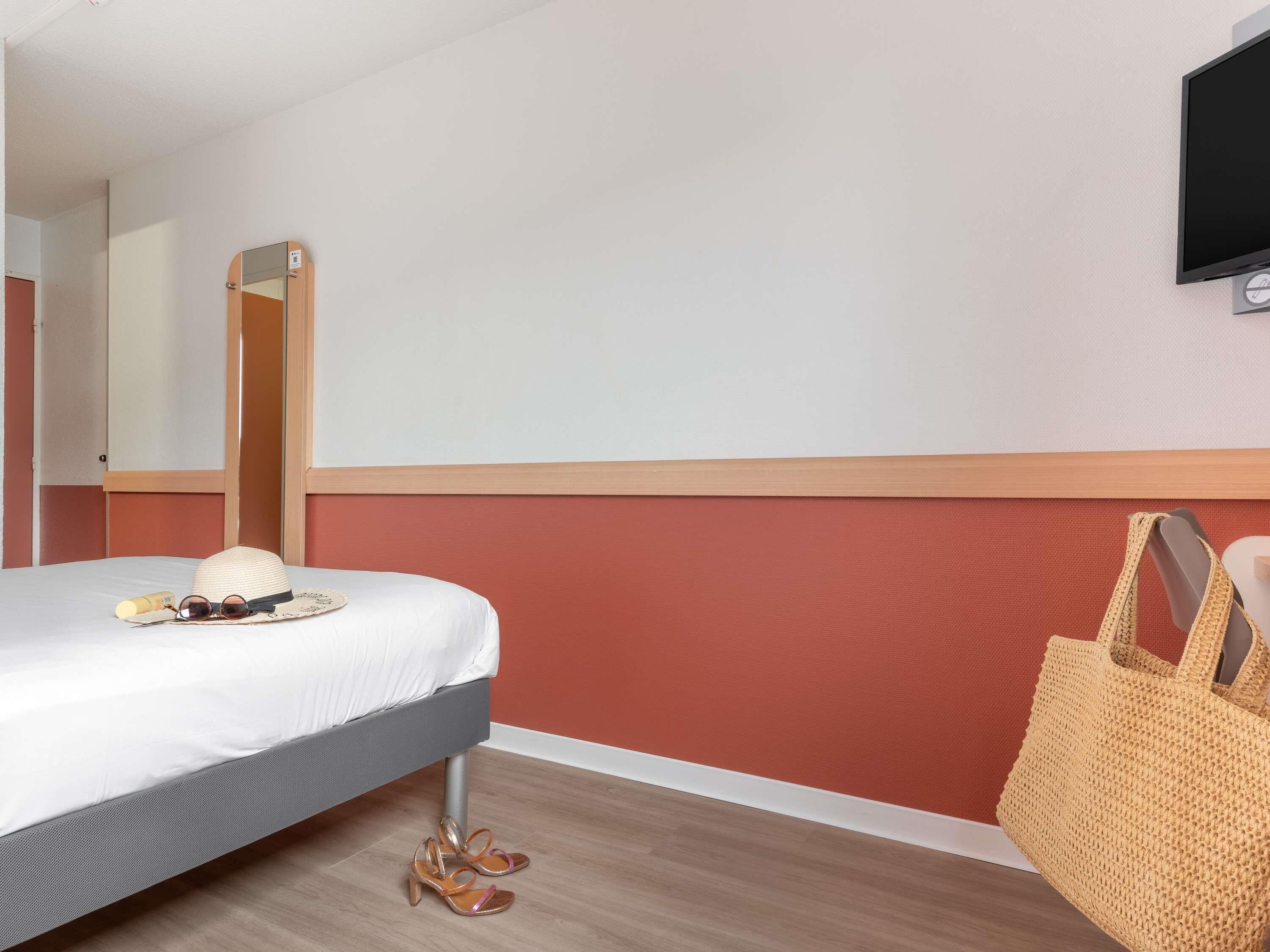 Ett hotellrum med en säng, en tv som är monterad på väggen, ett trägolv och en vävd väska som ligger på golvet.