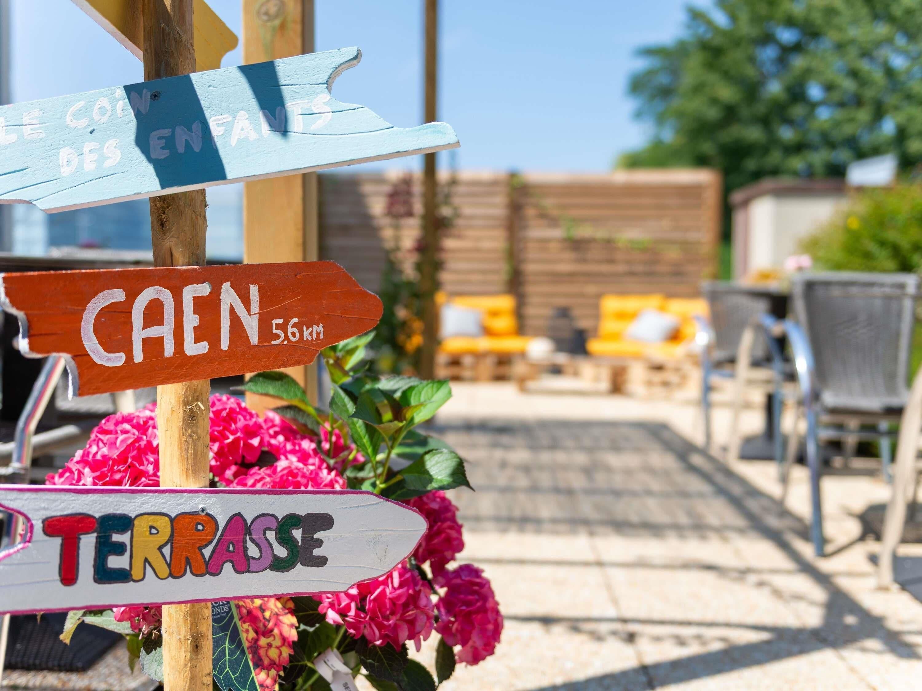 A wooden signpost with directional arrows pointing to different locations, including a café, a terrace, and a place called 'CAEN' with a distance of 5.6 km.