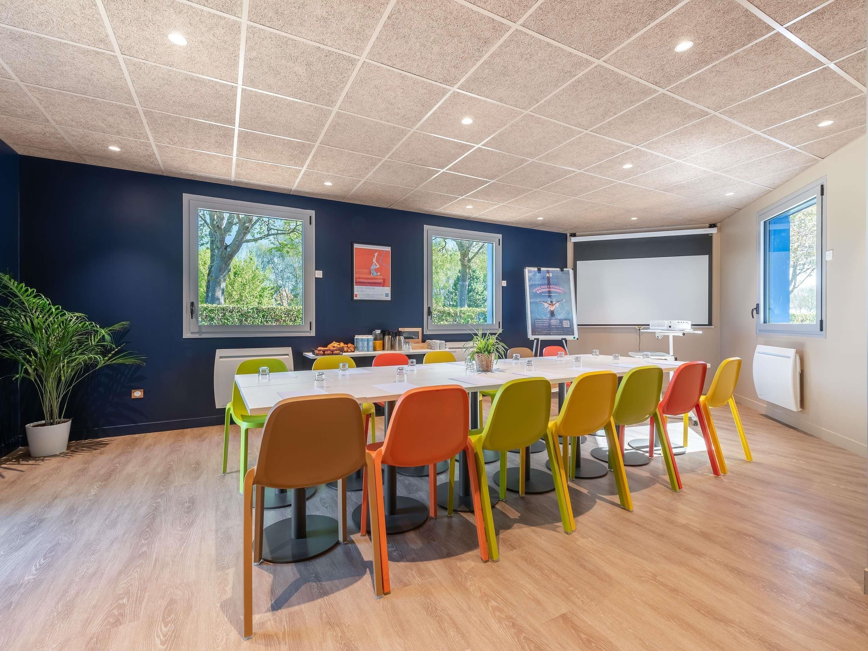 A conference room with a white table, colorful chairs, a projector screen, and a large window with a view of trees.