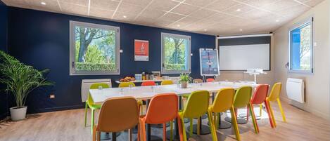 A conference room with a white table, colorful chairs, a projector screen, and a large window with a view of trees.