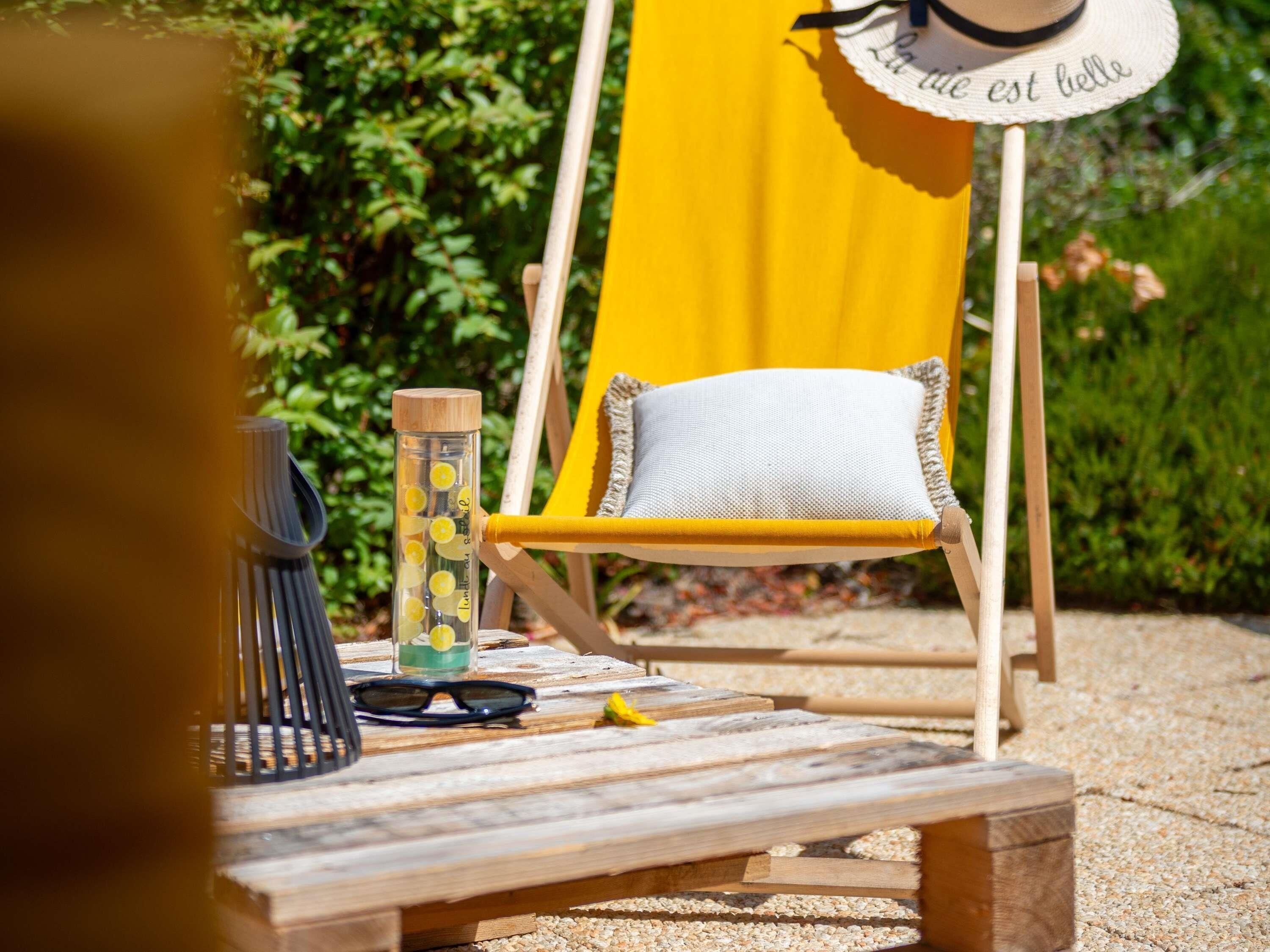 A wooden deck with a yellow chair, a clear bottle with yellow objects, a pair of sunglasses, and a small wooden block.