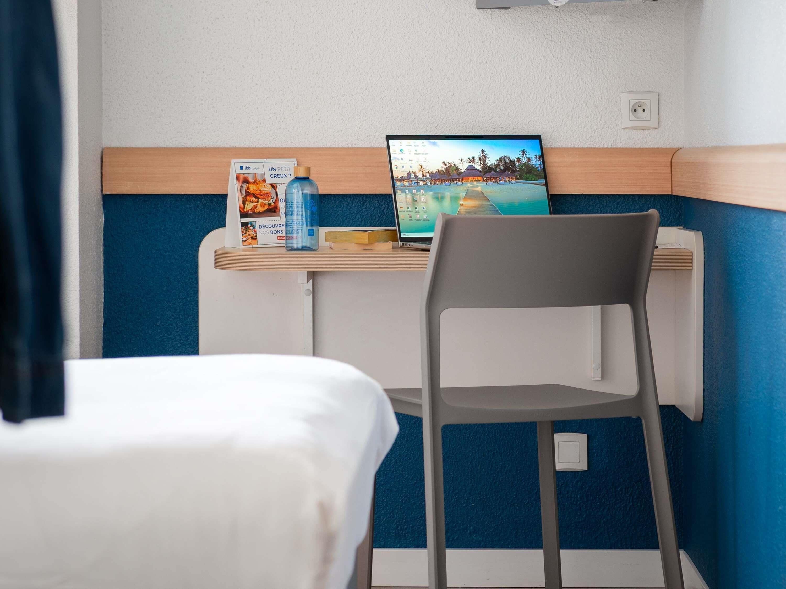 Une chambre d’hôtel avec un lit, un bureau avec un ordinateur portable, une chaise, une bouteille d’eau et une brochure posée sur le bureau.