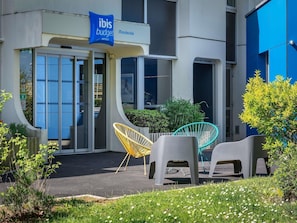 A hotel entrance with a sign for ibis budget hotels, outdoor seating with chairs, and a small garden area. - ibis Budget Caen Mondeville (Mondeville)