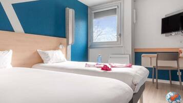 Quarto de hotel com duas camas, uma escrivaninha, uma cadeira, uma televisão e uma janela com vista para árvores.