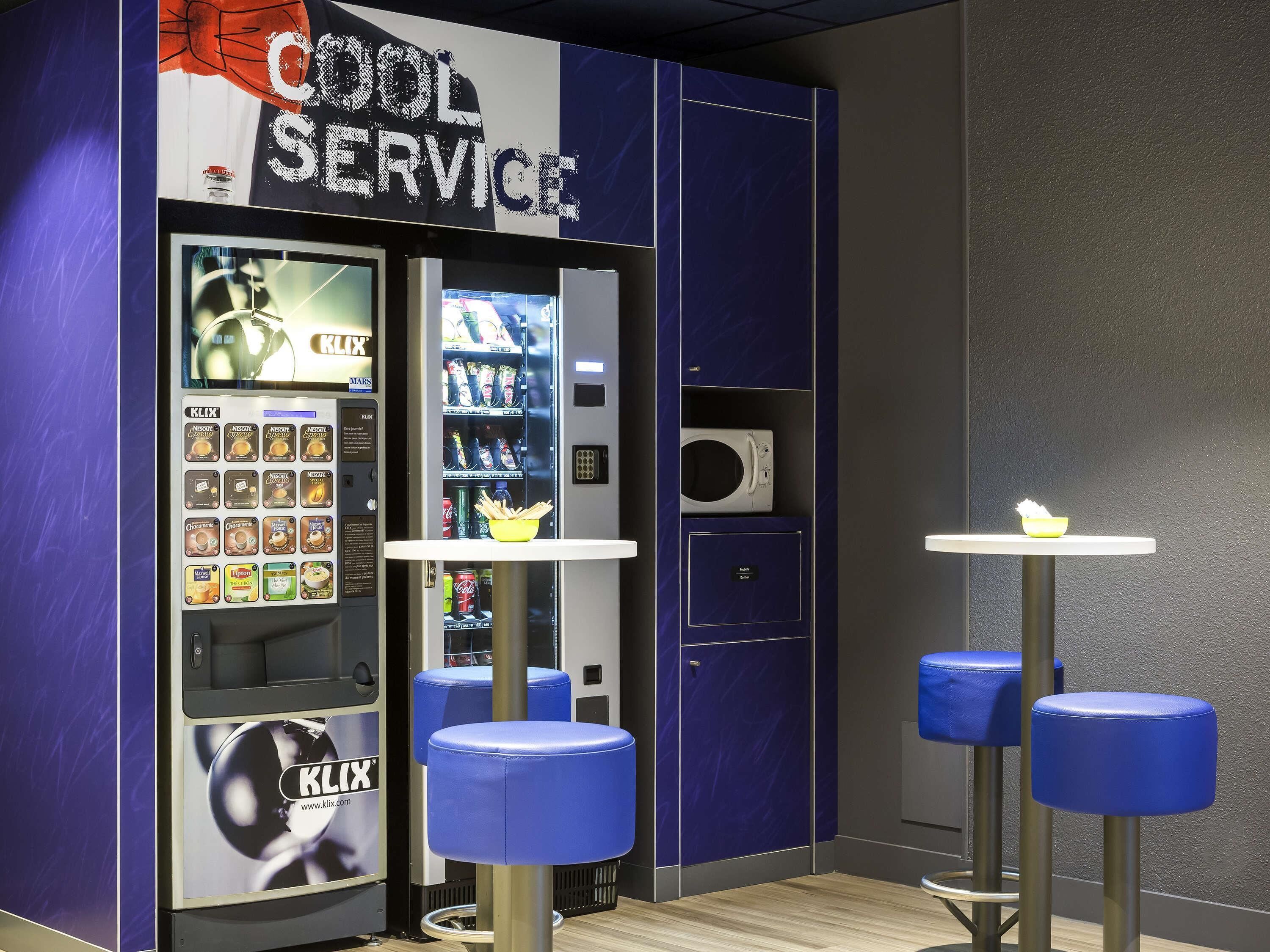 Una máquina expendedora con un rótulo que dice 'Cool Service', un microondas y taburetes azules.