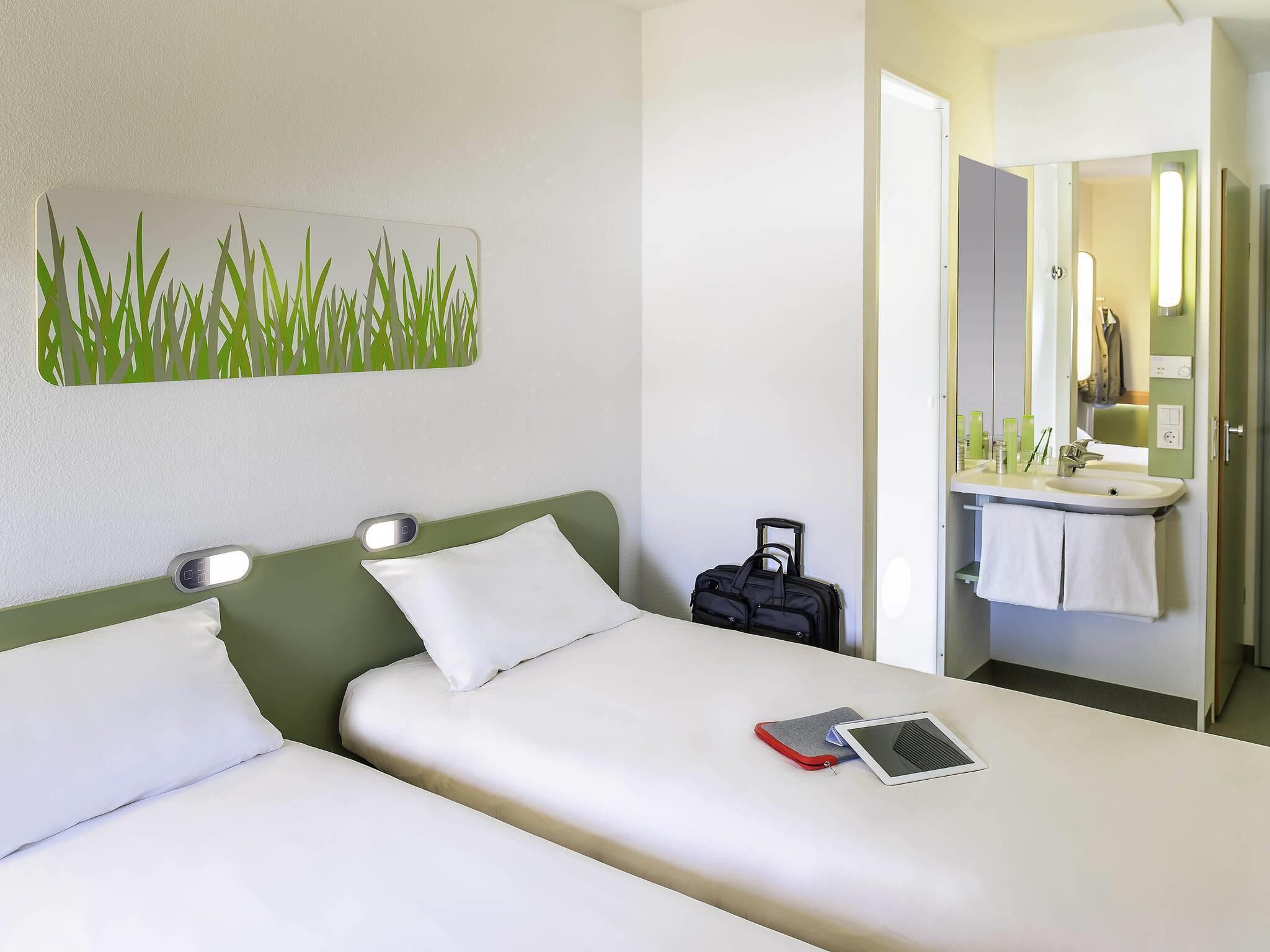 Une chambre d’hôtel comprenant un lit, une table de chevet, une valise, une salle de bain avec un lavabo et un miroir, et une œuvre d’art murale.