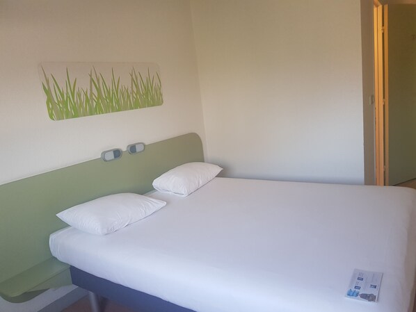 Una cama individual con ropa de cama blanca, un cabecero verde y una lámpara fijada a la pared.