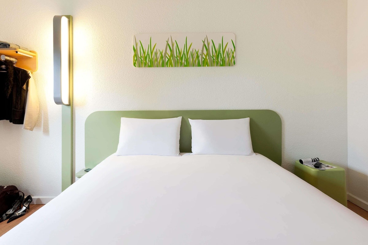 Een net opgemaakt bed met witte lakens, een groen hoofdbord en een aan de muur bevestigd kunstwerk van gras.