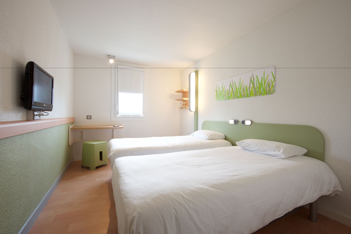 Une chambre d’hôtel avec deux lits, un petit tabouret, une télévision et une fenêtre.