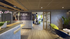 Een moderne receptiezone met een houten receptiebalie, een glazen scheidingswand en een zithoek met een blauwe stoel en een gele stoel.