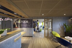 Un espace d’accueil moderne, doté d’un comptoir de réception en bois, d’une cloison en verre et d’un coin salon avec un fauteuil bleu et un fauteuil jaune.