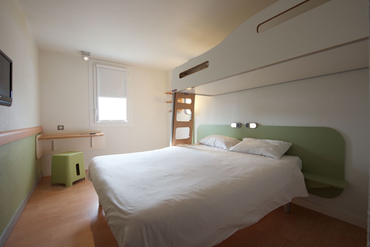 Un lit bien fait, avec une tête de lit verte et une literie blanche, dans une chambre simple et épurée, agrémentée d’un petit tabouret et d’une fenêtre.
