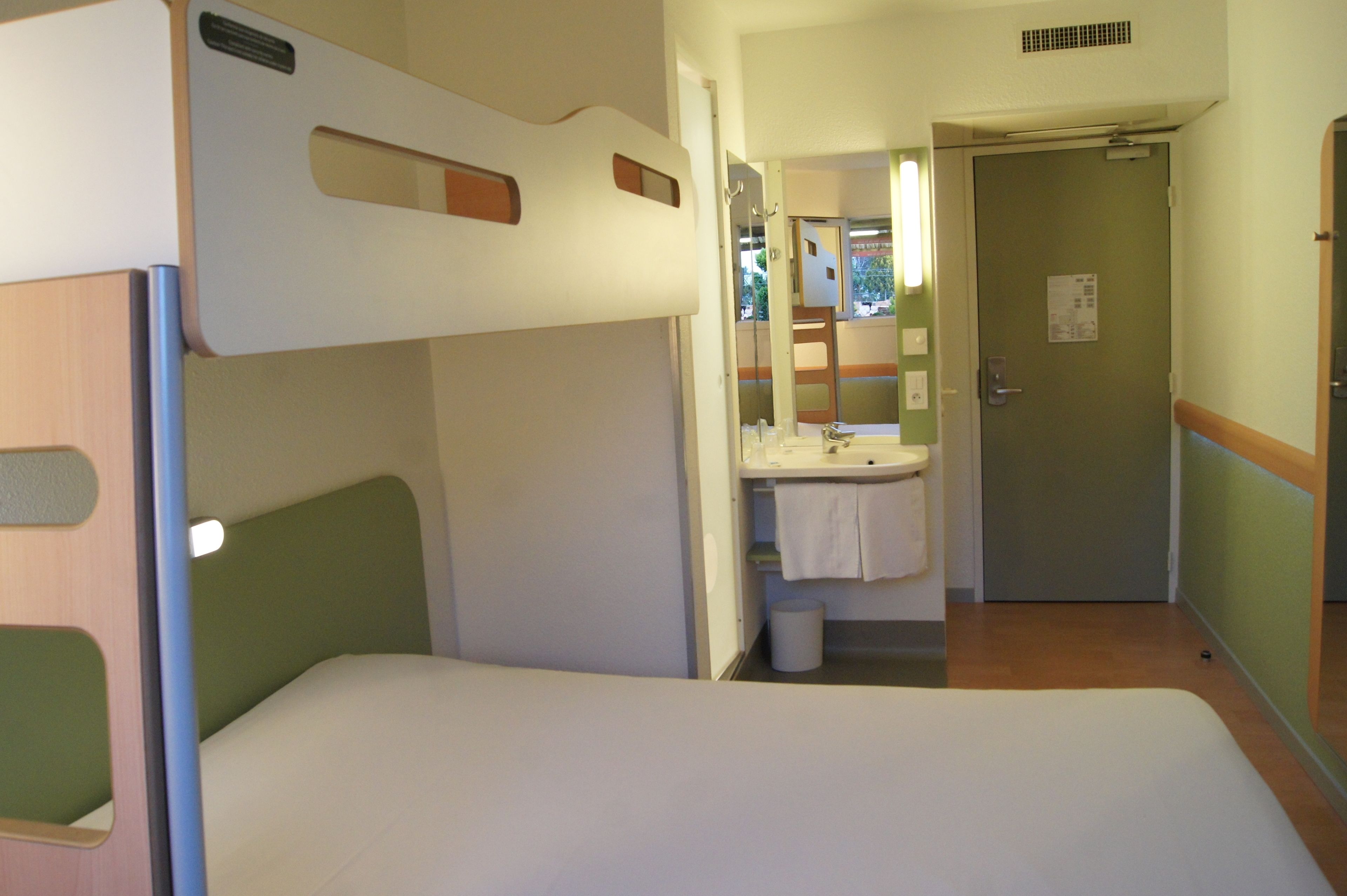 A compact hotel room with a bunk bed, a small bathroom with a sink and mirror, and a door leading to another room.