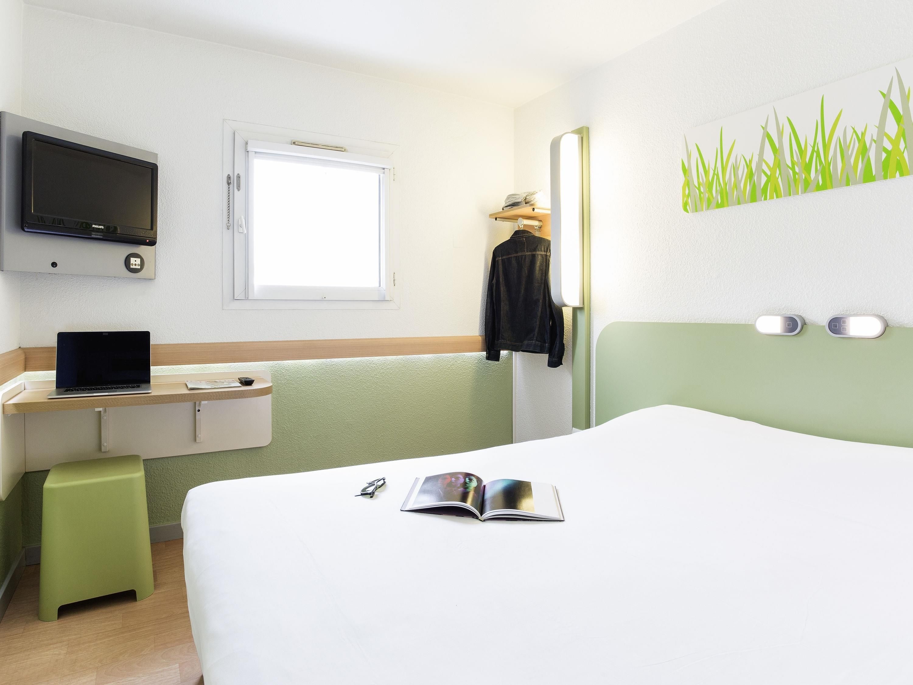 Ein Hotelzimmer mit einem Bett, einem Schreibtisch mit Laptop, einem an der Wand befestigten Fernseher, einem Fenster und einem Wandspiegel.