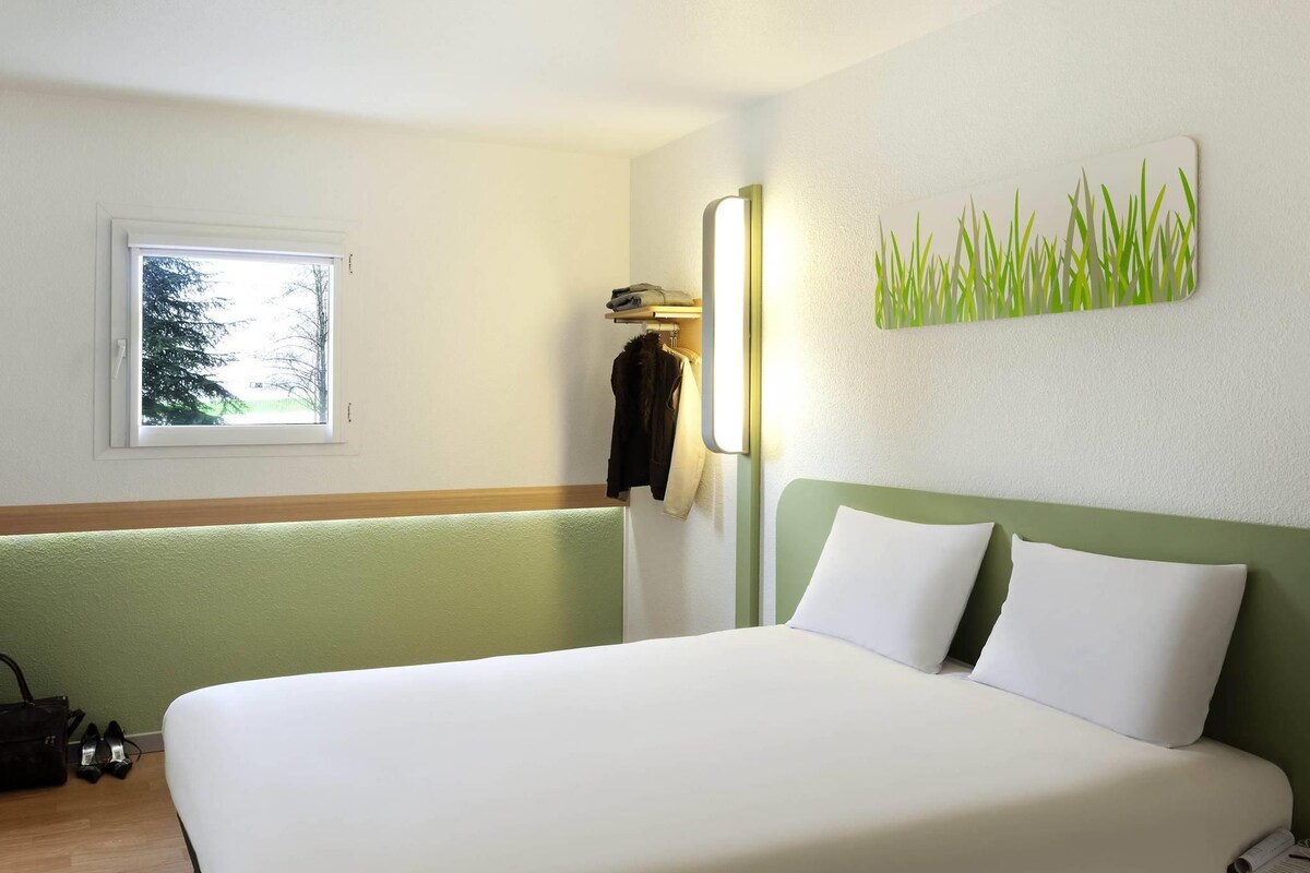 Habitación de hotel con cama, ventana, perchero y una lámpara fijada a la pared.