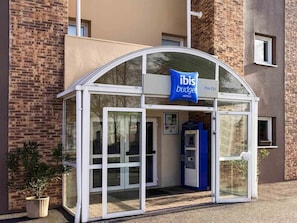 Ein moderner Hotelzugang mit Glasüberdachung, einem blauen Schild und einem Selbstbedienungskiosk.