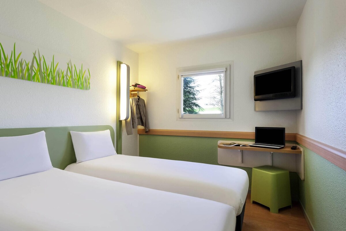 Habitación de hotel con dos camas, un escritorio con una computadora portátil, un televisor y una ventana con vista a árboles.