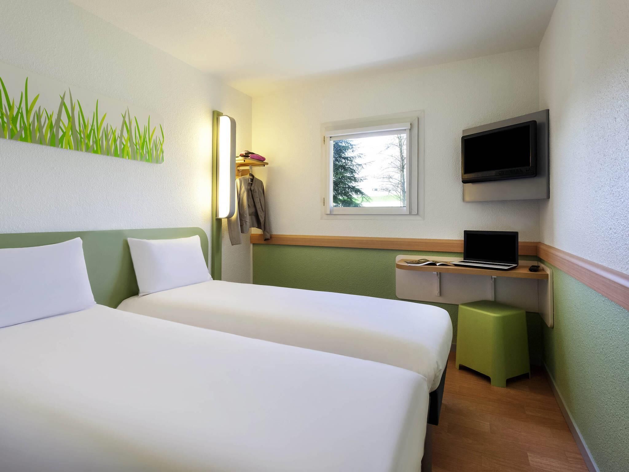 A hotel room with two beds, a desk with a laptop, a TV, and a window with a view of trees.