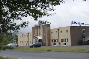 Een hotel met modern design, met een opvallend ‘ibis budget’ bord, een parkeerplaats en een auto ervoor geparkeerd.